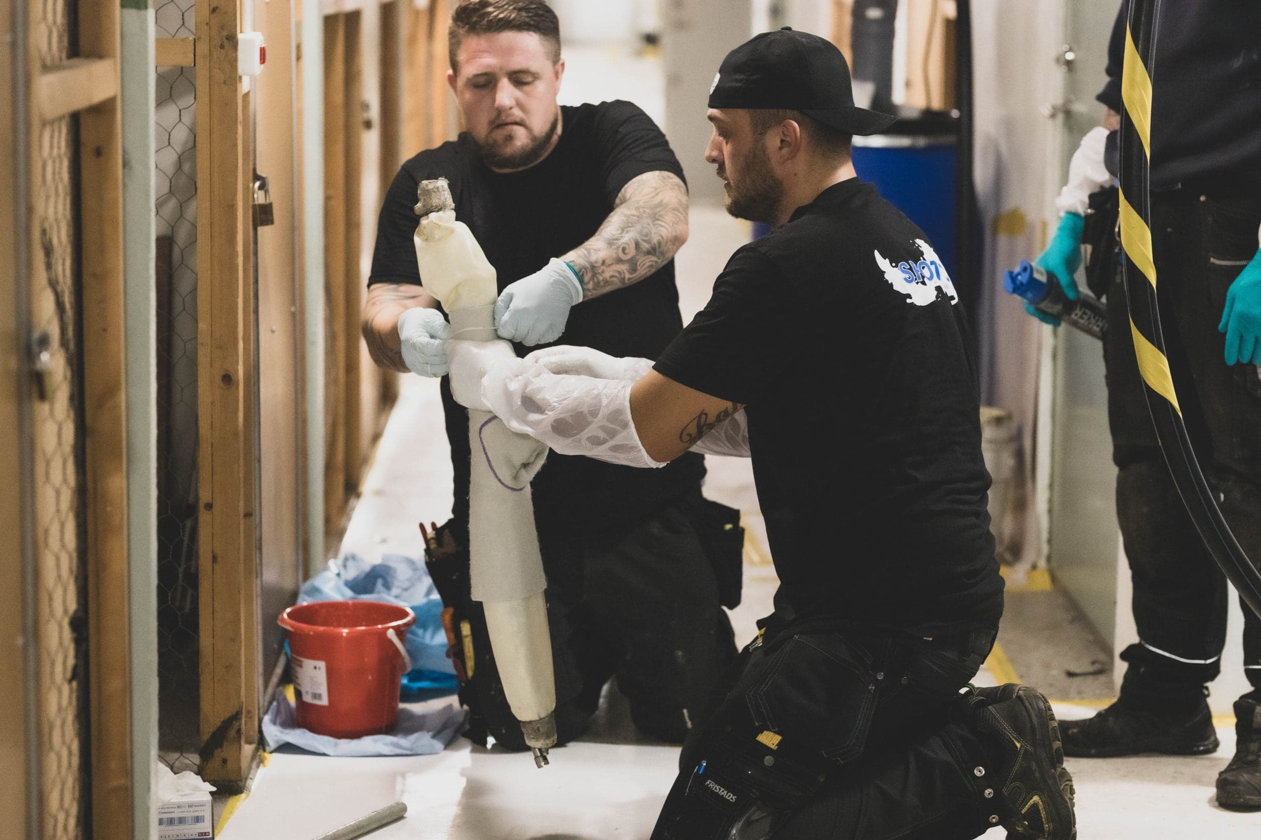 Två arbetare monterar Spoltec-isoleringshylsor på rör i en byggnadskorridor.